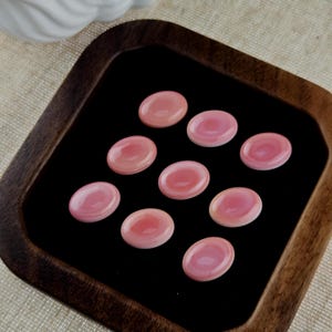 May include: Eight smooth, oval-shaped pink cabochons arranged in a grid pattern on a black velvet display tray. Each gemstone features a polished, glossy surface with subtle variations in light and dark pink tones, showcasing a rounded, domed profile.