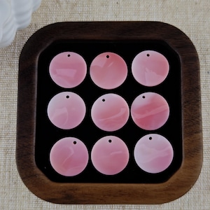 May include: Nine circular, light pink shell pendants arranged in a three-by-three grid. Each flat, smooth disc features a single small hole near the top edge for threading, showcasing a subtle, natural pearlescent sheen across the surface.