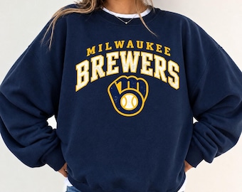 May include: A navy blue crewneck sweatshirt featuring the text MILWAUKEE BREWERS in a distressed yellow font. Below the text is a yellow graphic of a baseball glove holding a baseball, also with a distressed texture finish.