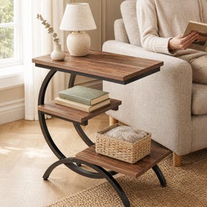 Peut inclure: Une table d'appoint en forme de C avec un cadre en métal foncé et trois étagères en bois. L'étagère supérieure porte une lampe beige et un petit vase avec de l'eucalyptus. L'étagère centrale contient deux livres et celle du bas un panier.