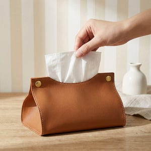Peut inclure: Un étui à mouchoirs en cuir beige avec une ouverture supérieure incurvée et deux boutons-pression en métal doré sur les coins supérieurs. Le support rectangulaire contient un mouchoir en papier blanc tiré par le centre.