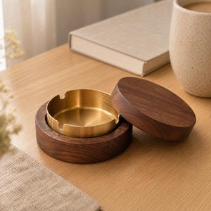 Può includere: Un posacenere rotondo in legno con finitura a venature di noce marrone scuro e un inserto rimovibile in ottone lucido. Il posacenere include un coperchio circolare in legno abbinato che poggia accanto alla base, rivelando l'interno metallico.