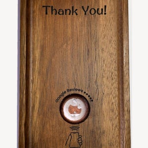 Puede incluir: Una bandeja rectangular de madera con esquinas redondeadas que presenta el texto Thank You! en la parte superior. En el centro hay una etiqueta NFC circular con el texto Google Reviews y cinco estrellas. Debajo, un icono de teléfono y el texto Tap your phone to support us.