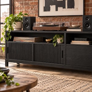 May include: A black media console featuring two sliding doors with vertical slats, one solid cabinet door, and an open top shelf. The unit is constructed with a dark wood grain finish and stands on four short legs, providing storage for electronics and media.