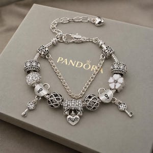 Könnte beinhalten: Ein silbernes Bettelarmband mit verschiedenen Perlen, darunter klare Kristalle, weiße Emaille-Blumen und herzförmige Schlösser. Ein zentraler Anhänger ist als Schleife gestaltet, mit dem Wort LOVE auf einem herzförmigen Anhänger eingraviert.