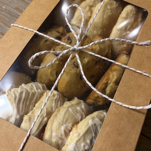 May include: A rectangular brown cardboard box with a clear plastic window containing an assortment of baked goods, including chocolate chip cookies and white-iced pastries, tied with a thin white and brown decorative twine bow.