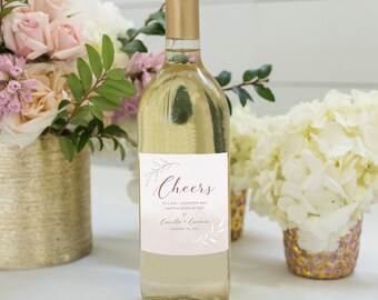 Puede incluir: Una botella de vino transparente con tapón dorado y etiqueta con la inscripción "Cheers". La etiqueta incluye los nombres Camilla & Cameron y la fecha 10 de enero de 2023. La botella está rodeada de arreglos florales.
