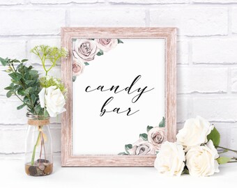May include: A framed sign with the words "candy bar" in elegant script. The sign is decorated with watercolor roses in the corners and is displayed on a white surface with floral arrangements and a white brick background.