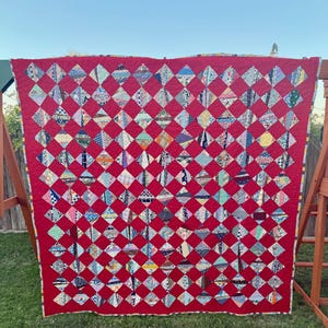 May include: A large rectangular quilt featuring a vibrant red background with a repeating grid of diamond-shaped fabric patches. Each diamond displays a unique multi-colored patterned textile, creating a high-contrast geometric design.