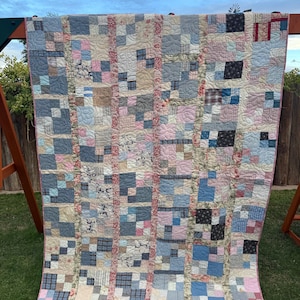 May include: A patchwork quilt featuring a grid of squares in various shades of blue, pink, and cream. The design incorporates diverse fabric patterns including stripes, plaids, and floral prints, separated by vertical strips of pink floral fabric.