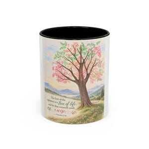 May include: A white ceramic mug with a black interior and rim, featuring a watercolor illustration of a tree with green and pink leaves in a landscape. The text reads: The fruit of the righteous is a tree of life; and he that winneth souls is wise. - Proverbs 11:30.