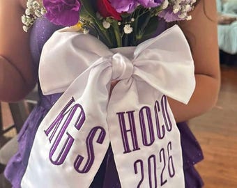 May include: A floral bouquet featuring deep red roses, vibrant purple carnations, and small white baby's breath flowers. A large white satin ribbon is tied around the stems, embroidered with purple text that reads KGS HOCO 2026.