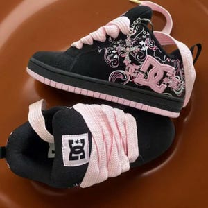 May include: A pair of black and light pink DC trainers featuring thick light pink laces, a prominent pink DC logo on the side, and a decorative silver-toned cross charm attached to the eyelets. The black upper displays a white and pink floral pattern.