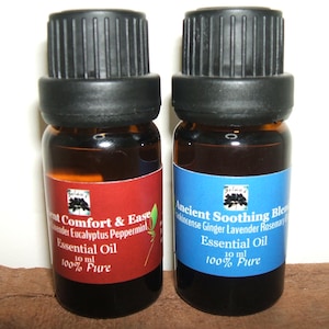 May include: Two small brown glass bottles with black caps and labels. The bottle on the left has a red label with the text "Comfort & Ease Lavender Eucalyptus Peppermint Essential Oil 10 ml 100% Pure". The bottle on the right has a blue label with the text "Ancient Soothing Blend Frankincense Ginger Lavender Rosemary Essential Oil 10 ml 100% Pure".