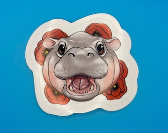 May include: A die-cut sticker featuring a grey hippopotamus head with a wide, open mouth showing teeth and a tongue. The hippo is surrounded by several vibrant red poppy flowers with dark centres, all set against a white border outline.