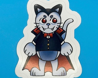 May include: A die-cut sticker featuring a light grey cat dressed as a vampire. The cat has red eyes, sharp fangs, and wears a black tuxedo jacket with a red bowtie and a long cape with a red interior lining. The sticker has a white border.