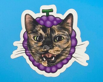 May include: A die-cut sticker featuring a tortoiseshell cat face framed by a cluster of purple grapes with a green stem. The cat has green eyes, long black whiskers, and an open mouth showing teeth, set against a solid bright blue background.