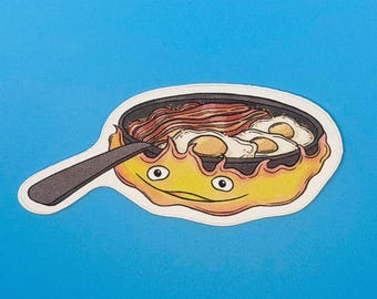 May include: A die-cut sticker featuring a black frying pan filled with cooked bacon and three fried eggs. The pan is held by a yellow and orange flame character with two eyes and a small mouth, creating a cartoon illustration.