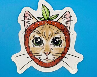 May include: A die-cut sticker featuring a tan cat with large green eyes wearing a red strawberry-shaped hood. The hood has a textured surface with small black dots and a green leaf at the top. The cat has visible whiskers and pink ears.