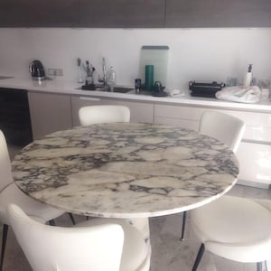 May include: A round dining table with a white and grey marble top featuring prominent dark veining, supported by a central pedestal base. Four white upholstered chairs with curved backrests and thin black metal legs surround the table.