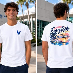 May include: A white short-sleeve t-shirt featuring a small blue eagle logo on the front left chest. The back displays a colorful graphic of a small airplane flying over palm trees and a sunset with the text Embry-Riddle Aeronautical University.
