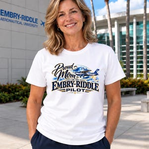 May include: A white short-sleeved t-shirt featuring a graphic print that reads Proud Mom of an Embry-Riddle Pilot, accompanied by an illustration of a small airplane flying over a globe with blue and gold accents.