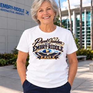 May include: A white short-sleeved t-shirt featuring a graphic print that reads Proud Grandma of an Embry-Riddle Eagle, accompanied by an illustration of a bald eagle, a globe, and a small heart icon in blue, gold, and black colors.