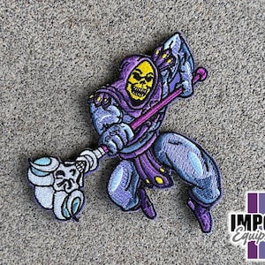 May include: Purple and yellow embroidered patch of Skeletor from Masters of the Universe, holding a staff with a skull on the end. The patch is on a gray background.