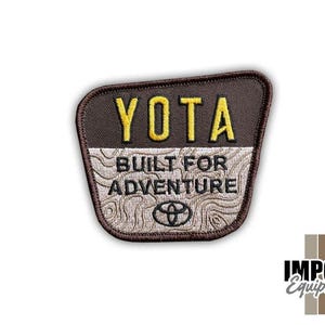 May include: A brown and tan embroidered patch with the word "YOTA" in yellow block letters. Below, the text reads "BUILT FOR ADVENTURE" above a Toyota logo. The patch has a textured design and a brown border.