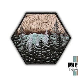 May include: Embroidered patch with a forest scene inside a hexagon. The design features dark green and gray pine trees, a light blue lake, and a brown mountain range under a beige sky. The patch has a black border.
