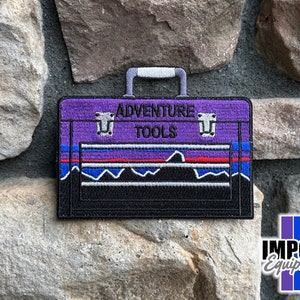 May include: Purple and black embroidered patch of a toolbox with the text "Adventure Tools" on the front. The patch has a blue, red, and white mountain range design.