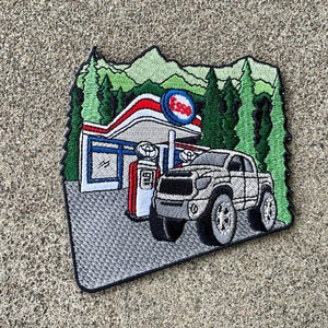 May include: Embroidered patch featuring an Esso gas station scene with a white truck. The patch includes green trees, mountains, and the Esso logo in red, white, and blue. The patch is gray and black.