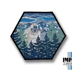May include: Embroidered patch in a hexagonal shape featuring a smiling yeti in a forest scene with mountains and trees. The patch has a black border and is predominantly blue, white, and green.