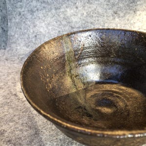May include: A dark brown ceramic bowl with a glossy, textured finish and a prominent vertical light-coloured glaze streak running from the rim towards the centre. The interior surface shows visible circular markings and a deep, reflective glaze.