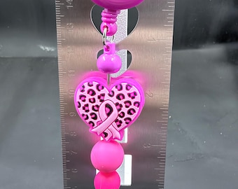Puede incluir: Un portacredenciales rosa brillante con un dije en forma de corazón con estampado de leopardo rosa y un lazo de concienciación rosa. El objeto se muestra frente a una regla metálica, con una longitud total de unos 14 cm.
