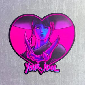 Puede incluir: Una pegatina con una ilustración estilizada de una persona con piel azul y ojos amarillos, sobre un corazón rosa brillante. Los brazos de la persona están cruzados y el texto "Your Idol" está escrito en una fuente estilizada.