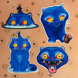 Peut inclure: Quatre autocollants de chats bleus avec des yeux jaunes et des bouches ouvertes. Un chat a une petite figurine sur la tête. Un autre chat tient un petit pot de fleurs. Les autocollants sont sur un fond rose avec des motifs dorés.