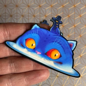 Puede incluir: Una pegatina de gato azul con ojos naranjas y un sombrero negro. El gato tiene un borde blanco y una nariz y orejas rosas. La pegatina se sostiene en una mano.