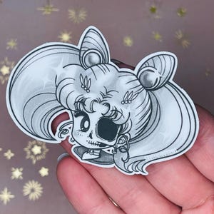 Könnte beinhalten: Schwarz-weißer Aufkleber mit einer Comicfigur mit Totenkopfgesicht und Zöpfen. Die Figur trägt eine Halskette und Ohrringe.