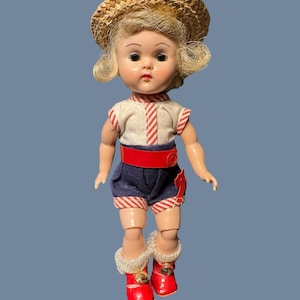 May include: A vintage doll with blonde hair wearing a straw hat, a white top with red stripes, navy blue shorts with red and white trim, a red belt, and red boots with white fur trim. The doll has a painted face with large eyes.