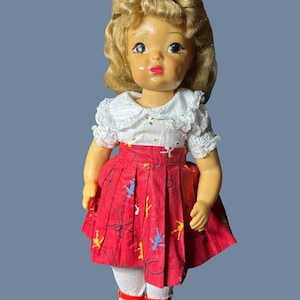 May include: A vintage doll with blonde hair, wearing a white blouse with a lace collar and a red skirt with a colorful pattern. The doll has red shoes and white socks. The doll is a collectible item.