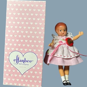 Könnte beinhalten: Eine Vintage-Effanbee-Puppe in einem rosa Kleid mit rotem Knopf, weißen Socken und schwarzen Schuhen. Die Puppe hat rotes Haar und steht neben einer rosa Schachtel mit einem Herzmuster und dem Effanbee-Logo.
