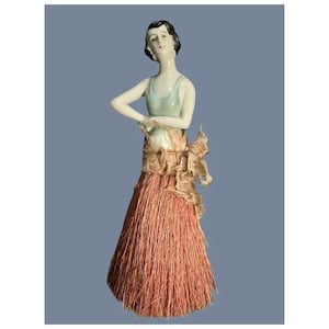 Puede incluir: Figura de porcelana de una mujer con un vestido estilo flapper. La figura tiene un top verde pálido, una cintura de color beige y dorado, y una falda larga de color marrón rojizo. La mujer tiene el pelo oscuro y posa con un brazo doblado.