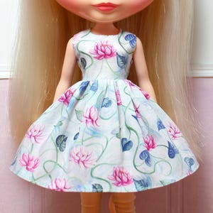 May include: A doll wearing a sleeveless dress with a floral pattern. The dress features pink lotus flowers, blue leaves, and green vines on a white background. The doll has blonde hair and is wearing light-colored tights.