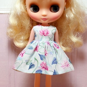 May include: A doll wearing a white dress with a floral pattern of pink lotus flowers and blue leaves. The doll has blonde hair and large blue eyes. The dress is sleeveless and has a pleated skirt.