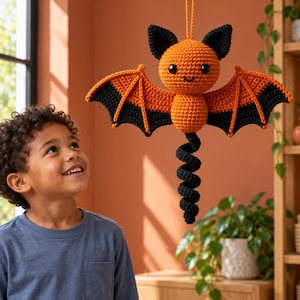 May include: A crocheted bat ornament featuring a round orange head and body, black ears, and black-tipped orange wings. The bat has a long, black, spiraled tail and a hanging loop at the top. The design includes small black eyes and a stitched smile.