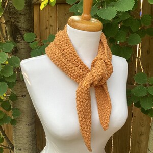 May include: A crocheted scarf in a warm orange colour, tied around a white mannequin's neck. The scarf is made with a simple stitch and has a loose, flowing texture.