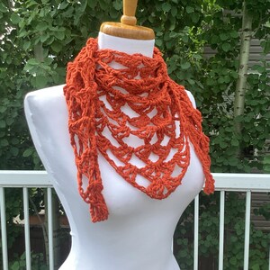 May include: A crocheted orange scarf with a triangular shape and fringe details. The scarf is displayed on a white mannequin.