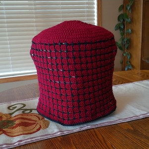May include: A handmade, red crochet tea cosy with a black grid pattern. The cosy is designed to fit over a teapot and keep the tea warm. The cosy is sitting on a wooden table.