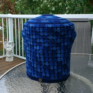 May include: A handmade, blue crochet cover in various shades of blue. The cover is cylindrical with a rounded top and a textured, open weave pattern. A small, rectangular tag is attached to the bottom.